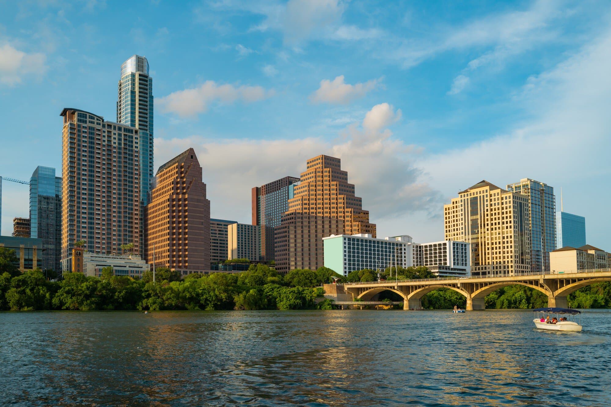 A city skyline viewed across a wide river with an arched bridge and a small boat in the water.