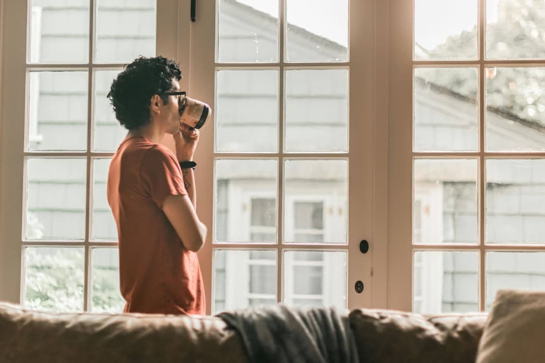 Man Looking Out Window At House Drinking Coffee